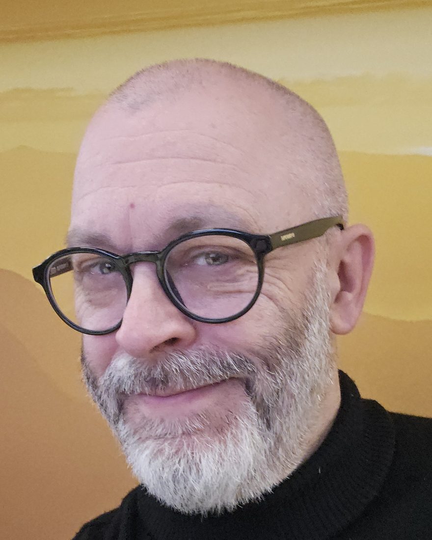Dominic Townsend, smiling, wearing round black glasses and a black roll-neck